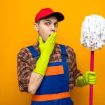 scared covered mouth with hand young cleaning guy wearing uniform and cap with gloves holding mop isolated on orange background