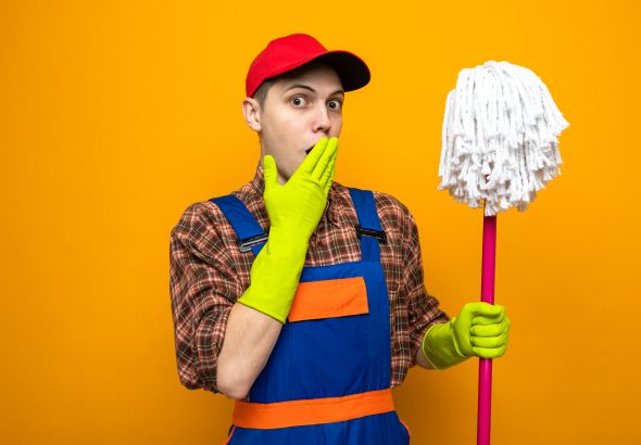 scared covered mouth with hand young cleaning guy wearing uniform and cap with gloves holding mop isolated on orange background