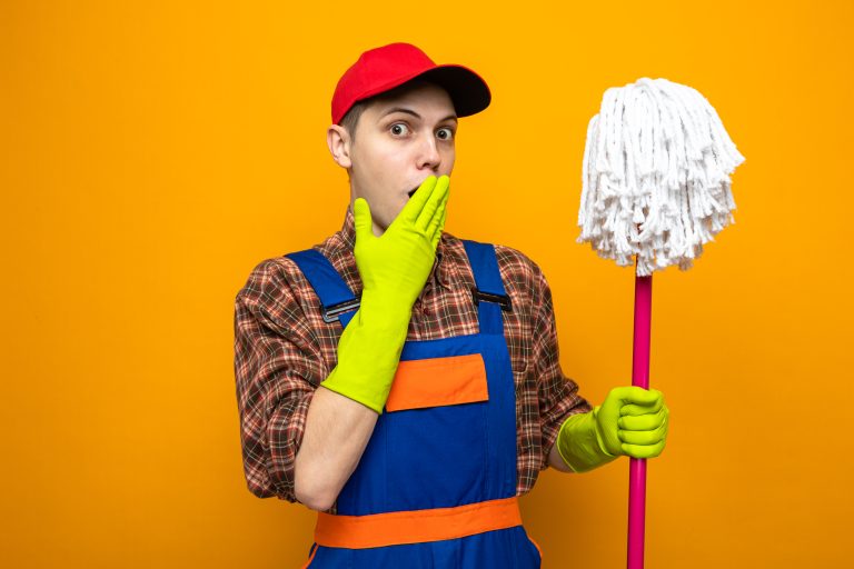 scared covered mouth with hand young cleaning guy wearing uniform and cap with gloves holding mop isolated on orange background